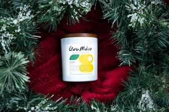 A candle with a white label and a brown lid is placed on a bright red fabric, surrounded by green pine branches with white snow-like accents. The candle label has modern typography and minimalistic design elements.
