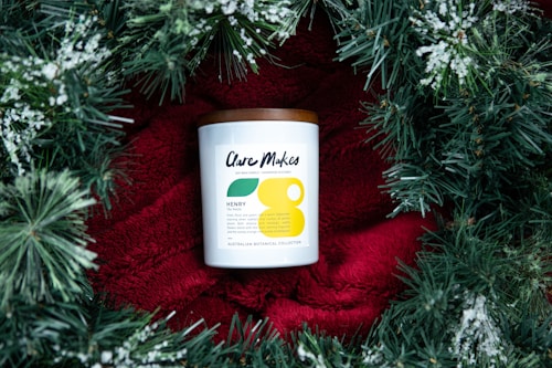 A candle with a white label and a brown lid is placed on a bright red fabric, surrounded by green pine branches with white snow-like accents. The candle label has modern typography and minimalistic design elements.