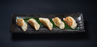 Five pan-fried dumplings are neatly arranged on a long, rectangular, dark ceramic plate. A green leaf lies beneath the dumplings, adding a contrasting element to the presentation. The dumplings have a golden-brown, crispy side while the top is a lighter, steamed texture.