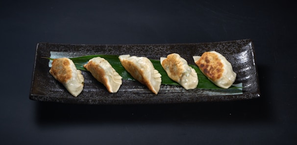 Five pan-fried dumplings are neatly arranged on a long, rectangular, dark ceramic plate. A green leaf lies beneath the dumplings, adding a contrasting element to the presentation. The dumplings have a golden-brown, crispy side while the top is a lighter, steamed texture.