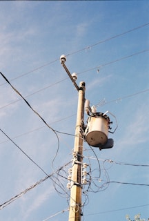 A utility pole with multiple electrical wires extending outward against a clear blue sky. At the top of the pole, there is a transformer and several insulators holding the wires in place.
