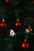 Green Christmas tree branches adorned with shiny red baubles, a white ornament in the shape of a tree with a red reindeer design, and a small figurine of an animal.
