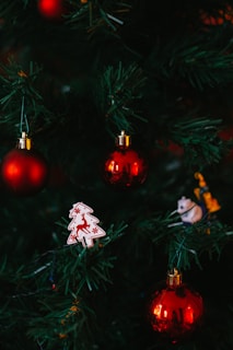 Green Christmas tree branches adorned with shiny red baubles, a white ornament in the shape of a tree with a red reindeer design, and a small figurine of an animal.