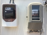 Two electricity meters mounted on a wall. The meter on the left is housed in a white and brown case with a digital display, while the meter on the right is older, encased in a beige cover with a glass panel showing the numeric readings.