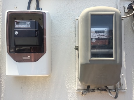 Two electricity meters mounted on a wall. The meter on the left is housed in a white and brown case with a digital display, while the meter on the right is older, encased in a beige cover with a glass panel showing the numeric readings.