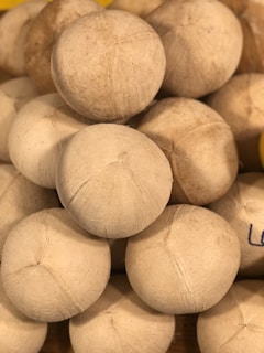 A stack of peeled coconuts with a rough, fibrous texture. The coconuts are arranged in a pyramid shape, closely packed together.
