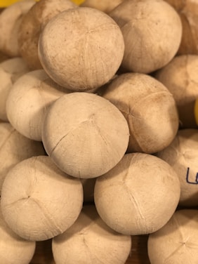 A stack of peeled coconuts with a rough, fibrous texture. The coconuts are arranged in a pyramid shape, closely packed together.