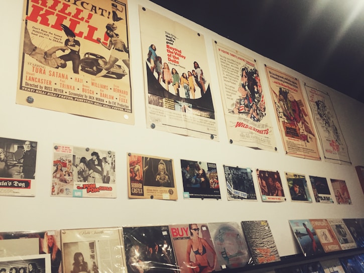 A wall display featuring vintage movie posters and album covers. The posters are arranged in two horizontal rows, with the top row consisting of large movie posters showcasing various films, some with retro themes. Below them are smaller images, likely related to films or music, and a series of vinyl records or album covers displayed on a shelf. The overall aesthetic is nostalgic, reminiscent of mid-20th century pop culture.