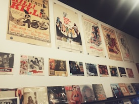 A wall display featuring vintage movie posters and album covers. The posters are arranged in two horizontal rows, with the top row consisting of large movie posters showcasing various films, some with retro themes. Below them are smaller images, likely related to films or music, and a series of vinyl records or album covers displayed on a shelf. The overall aesthetic is nostalgic, reminiscent of mid-20th century pop culture.