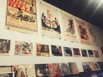 A wall display featuring vintage movie posters and album covers. The posters are arranged in two horizontal rows, with the top row consisting of large movie posters showcasing various films, some with retro themes. Below them are smaller images, likely related to films or music, and a series of vinyl records or album covers displayed on a shelf. The overall aesthetic is nostalgic, reminiscent of mid-20th century pop culture.
