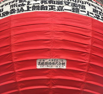 A large red lantern with horizontal ridges and Japanese characters in black printed in horizontal lines on a white background near the top. A label with more Japanese text is visible on a lower part of the lantern.