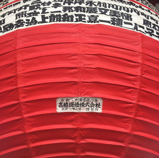 A large red lantern with horizontal ridges and Japanese characters in black printed in horizontal lines on a white background near the top. A label with more Japanese text is visible on a lower part of the lantern.