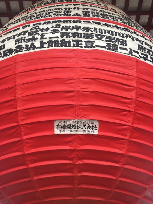A large red lantern with horizontal ridges and Japanese characters in black printed in horizontal lines on a white background near the top. A label with more Japanese text is visible on a lower part of the lantern.
