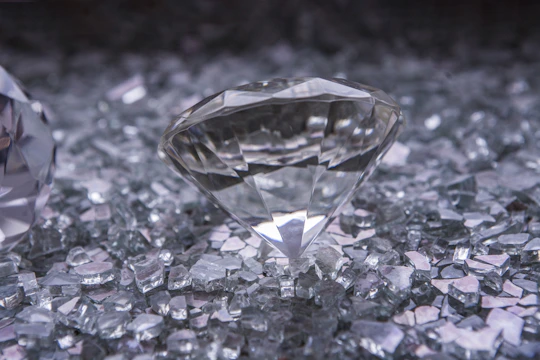 A large, clear diamond is placed on a surface covered with smaller sparkling crystals. The diamond's facets are sharply defined, and it reflects light brilliantly, creating a luminous effect. The surrounding crystals vary in size and enhance the overall shimmer.