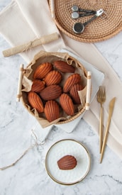 A basket filled with a rustic paper liner holding several baked madeleines is placed on a light-colored textured surface, possibly a marble countertop. To the top right of the basket, a round wicker tray holds a set of metal measuring spoons. Below the basket, on a woven beige cloth, are a gold-colored fork and knife. A single madeleine is placed on a white, textured plate with a gold rim near the bottom of the image. The setting has a clean and minimalistic arrangement.