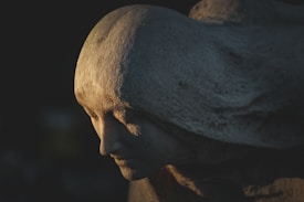 A stone sculpture of a serene face with gentle, flowing lines resembling hair or fabric. The play of light and shadow accentuates the calm expression, creating an ethereal and timeless feel.