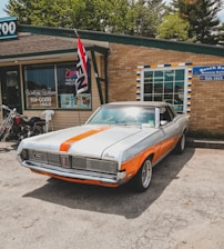 An orange and white vintage muscle car is parked in front of a brick building housing a beauty salon and a tattoo shop. A 'Open' flag is flying near the entrance, and a motorcycle is parked nearby. The car is well-maintained with polished chrome detailing and a soft top.