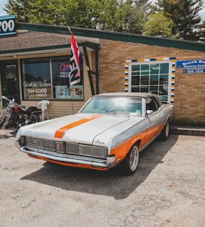 An orange and white vintage muscle car is parked in front of a brick building housing a beauty salon and a tattoo shop. A 'Open' flag is flying near the entrance, and a motorcycle is parked nearby. The car is well-maintained with polished chrome detailing and a soft top.