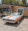 An orange and white vintage muscle car is parked in front of a brick building housing a beauty salon and a tattoo shop. A 'Open' flag is flying near the entrance, and a motorcycle is parked nearby. The car is well-maintained with polished chrome detailing and a soft top.