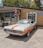 An orange and white vintage muscle car is parked in front of a brick building housing a beauty salon and a tattoo shop. A 'Open' flag is flying near the entrance, and a motorcycle is parked nearby. The car is well-maintained with polished chrome detailing and a soft top.