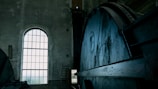 An industrial interior featuring a large, metal mechanism with gears and wheels taking up most of the view on the right. A tall arched window on the left allows light to filter into the space, highlighting the aged, concrete walls and the metal surfaces. The setting appears to be a factory or power plant, with a ladder visible in the background.