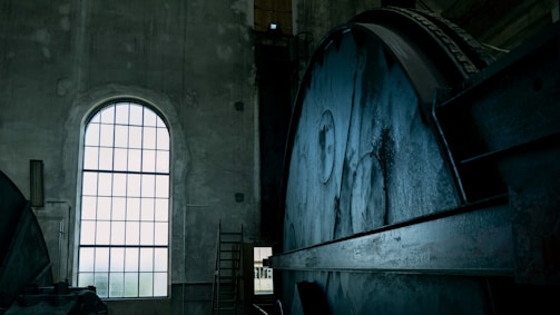 An industrial interior featuring a large, metal mechanism with gears and wheels taking up most of the view on the right. A tall arched window on the left allows light to filter into the space, highlighting the aged, concrete walls and the metal surfaces. The setting appears to be a factory or power plant, with a ladder visible in the background.