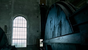 An industrial interior featuring a large, metal mechanism with gears and wheels taking up most of the view on the right. A tall arched window on the left allows light to filter into the space, highlighting the aged, concrete walls and the metal surfaces. The setting appears to be a factory or power plant, with a ladder visible in the background.