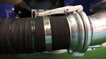 A close-up view of a heavy-duty metal pipe connection featuring a large rubber hose secured with metal clamps. The components display industrial material textures with a combination of metallic and rubber surfaces.