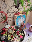A religious setup featuring a framed picture of a deity placed against a wall. There is a pot filled with soil and scattered with yellow and red flower petals. Incense sticks are burning, creating an aura of spirituality. A small bowl with liquid and some flowers is also placed in the pot. The surroundings include some additional small objects and a copper vessel placed nearby.