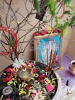 A religious setup featuring a framed picture of a deity placed against a wall. There is a pot filled with soil and scattered with yellow and red flower petals. Incense sticks are burning, creating an aura of spirituality. A small bowl with liquid and some flowers is also placed in the pot. The surroundings include some additional small objects and a copper vessel placed nearby.