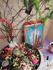 A religious setup featuring a framed picture of a deity placed against a wall. There is a pot filled with soil and scattered with yellow and red flower petals. Incense sticks are burning, creating an aura of spirituality. A small bowl with liquid and some flowers is also placed in the pot. The surroundings include some additional small objects and a copper vessel placed nearby.