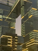 An architectural scene featuring modern skyscrapers with reflective glass facades and illuminated edges. The buildings have a geometric, stacked design with horizontal and vertical lines, creating a futuristic and sophisticated appearance. The sky, partially visible between the structures, adds contrast to the lit details of the architecture.