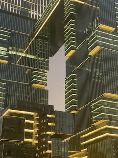An architectural scene featuring modern skyscrapers with reflective glass facades and illuminated edges. The buildings have a geometric, stacked design with horizontal and vertical lines, creating a futuristic and sophisticated appearance. The sky, partially visible between the structures, adds contrast to the lit details of the architecture.