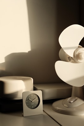 A minimalist scene featuring a simple analog clock with a dark face and sleek design sits on a table. To the right, there is a white fan with large blades casting soft shadows on the wall. A white, rounded electronic device, perhaps a speaker or humidifier, is also on the table. The lighting is warm, likely from natural sunlight coming through a window, creating a calm and serene atmosphere.