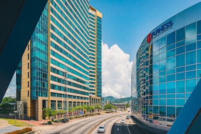 A modern urban scene featuring tall, glass-covered office buildings with reflective surfaces on either side of a curving road. The sky is clear with a few scattered clouds, and there are cars traveling along the road. The buildings have a sleek, corporate appearance with blue-tinted windows.