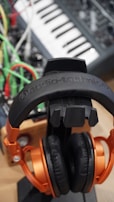 A pair of black and orange headphones rests on a stand. Behind them, a synthesizer or music keyboard is visible along with a tangle of bright colored cables in green, red, and yellow.