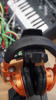 A pair of black and orange headphones rests on a stand. Behind them, a synthesizer or music keyboard is visible along with a tangle of bright colored cables in green, red, and yellow.