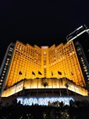 A grand hotel building illuminated with warm orange lights at night. The main facade features tall windows arranged in a geometric pattern and numerous flags flying in the front. The sign 'Plaza Indonesia' is prominently displayed in glowing letters above an entrance area surrounded by dark foliage.