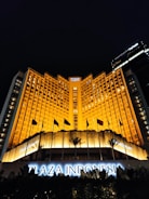 A grand hotel building illuminated with warm orange lights at night. The main facade features tall windows arranged in a geometric pattern and numerous flags flying in the front. The sign 'Plaza Indonesia' is prominently displayed in glowing letters above an entrance area surrounded by dark foliage.