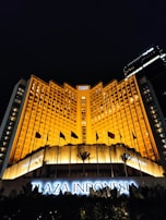A grand hotel building illuminated with warm orange lights at night. The main facade features tall windows arranged in a geometric pattern and numerous flags flying in the front. The sign 'Plaza Indonesia' is prominently displayed in glowing letters above an entrance area surrounded by dark foliage.