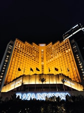 A grand hotel building illuminated with warm orange lights at night. The main facade features tall windows arranged in a geometric pattern and numerous flags flying in the front. The sign 'Plaza Indonesia' is prominently displayed in glowing letters above an entrance area surrounded by dark foliage.