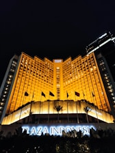 A grand hotel building illuminated with warm orange lights at night. The main facade features tall windows arranged in a geometric pattern and numerous flags flying in the front. The sign 'Plaza Indonesia' is prominently displayed in glowing letters above an entrance area surrounded by dark foliage.