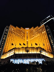 A grand hotel building illuminated with warm orange lights at night. The main facade features tall windows arranged in a geometric pattern and numerous flags flying in the front. The sign 'Plaza Indonesia' is prominently displayed in glowing letters above an entrance area surrounded by dark foliage.