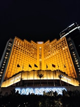 A grand hotel building illuminated with warm orange lights at night. The main facade features tall windows arranged in a geometric pattern and numerous flags flying in the front. The sign 'Plaza Indonesia' is prominently displayed in glowing letters above an entrance area surrounded by dark foliage.