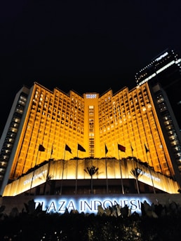 A grand hotel building illuminated with warm orange lights at night. The main facade features tall windows arranged in a geometric pattern and numerous flags flying in the front. The sign 'Plaza Indonesia' is prominently displayed in glowing letters above an entrance area surrounded by dark foliage.
