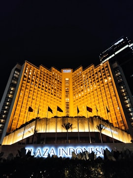 A grand hotel building illuminated with warm orange lights at night. The main facade features tall windows arranged in a geometric pattern and numerous flags flying in the front. The sign 'Plaza Indonesia' is prominently displayed in glowing letters above an entrance area surrounded by dark foliage.