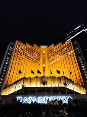 A grand hotel building illuminated with warm orange lights at night. The main facade features tall windows arranged in a geometric pattern and numerous flags flying in the front. The sign 'Plaza Indonesia' is prominently displayed in glowing letters above an entrance area surrounded by dark foliage.