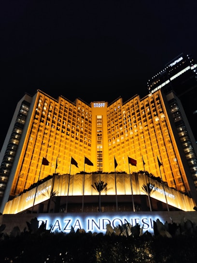 A grand hotel building illuminated with warm orange lights at night. The main facade features tall windows arranged in a geometric pattern and numerous flags flying in the front. The sign 'Plaza Indonesia' is prominently displayed in glowing letters above an entrance area surrounded by dark foliage.