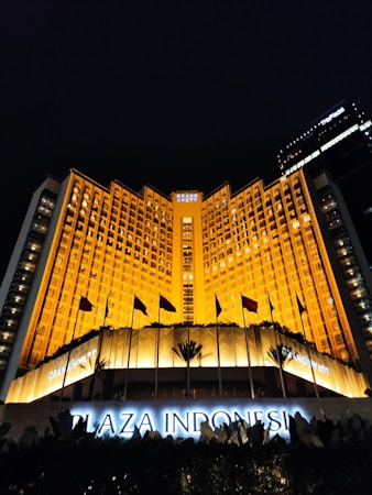A grand hotel building illuminated with warm orange lights at night. The main facade features tall windows arranged in a geometric pattern and numerous flags flying in the front. The sign 'Plaza Indonesia' is prominently displayed in glowing letters above an entrance area surrounded by dark foliage.