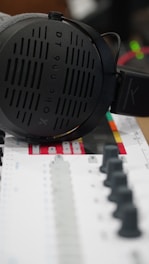A pair of large, over-ear headphones rests on a colorful mixing console. The headphones are sleek and modern, with a visible brand logo and model number. The mixing console features numerous faders and knobs, with a color scheme that includes black, gray, and red accents.